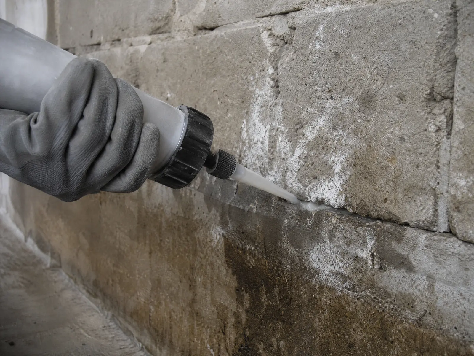 Une main injecte un produit d'étanchéité dans une fissure d'un vieux mur humide marqué par des remontées capillaires.