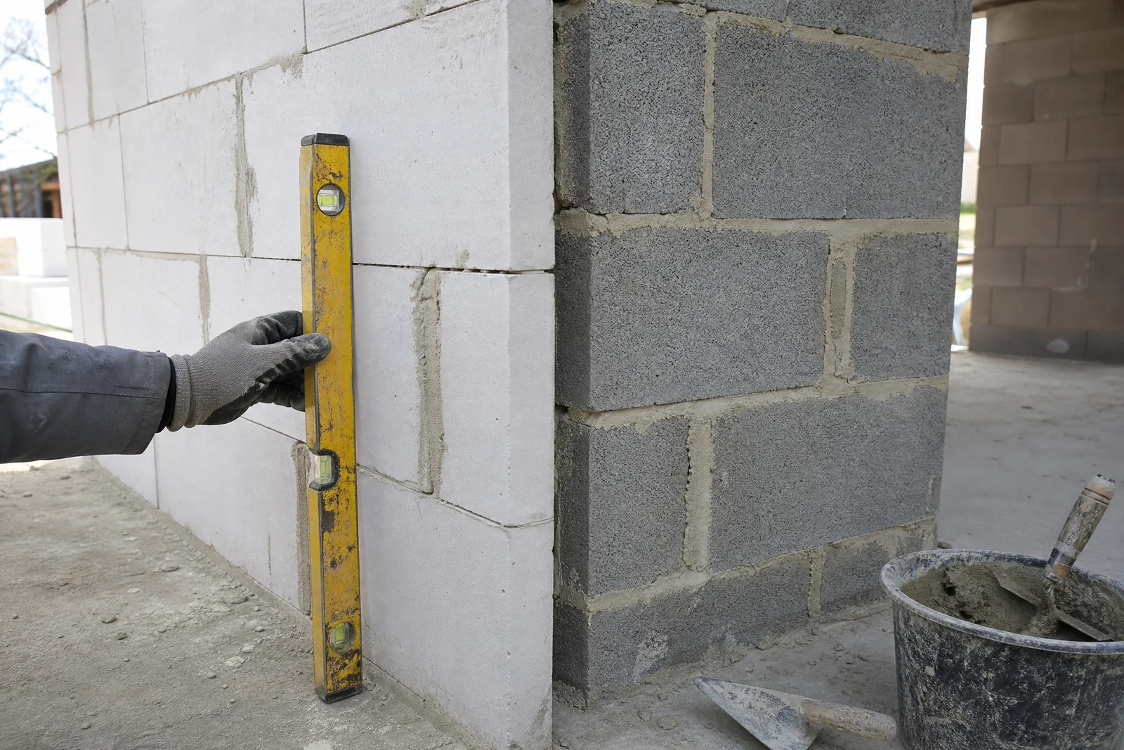 Main posant un niveau contre un mur en construction montrant côte à côte des blocs de béton cellulaire et des parpaings avec mortier frais.