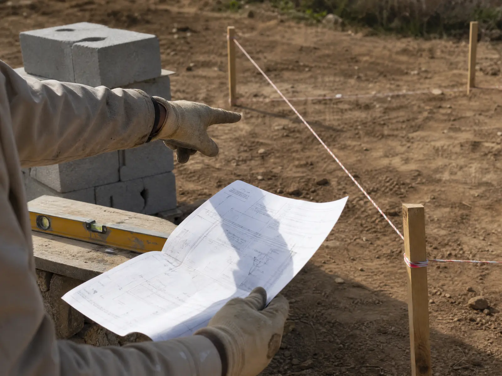 Main de maçon tenant un document administrative pointant les limites d'un terrain sur lequel sont empilés des parpaings et des outils de chantier.
