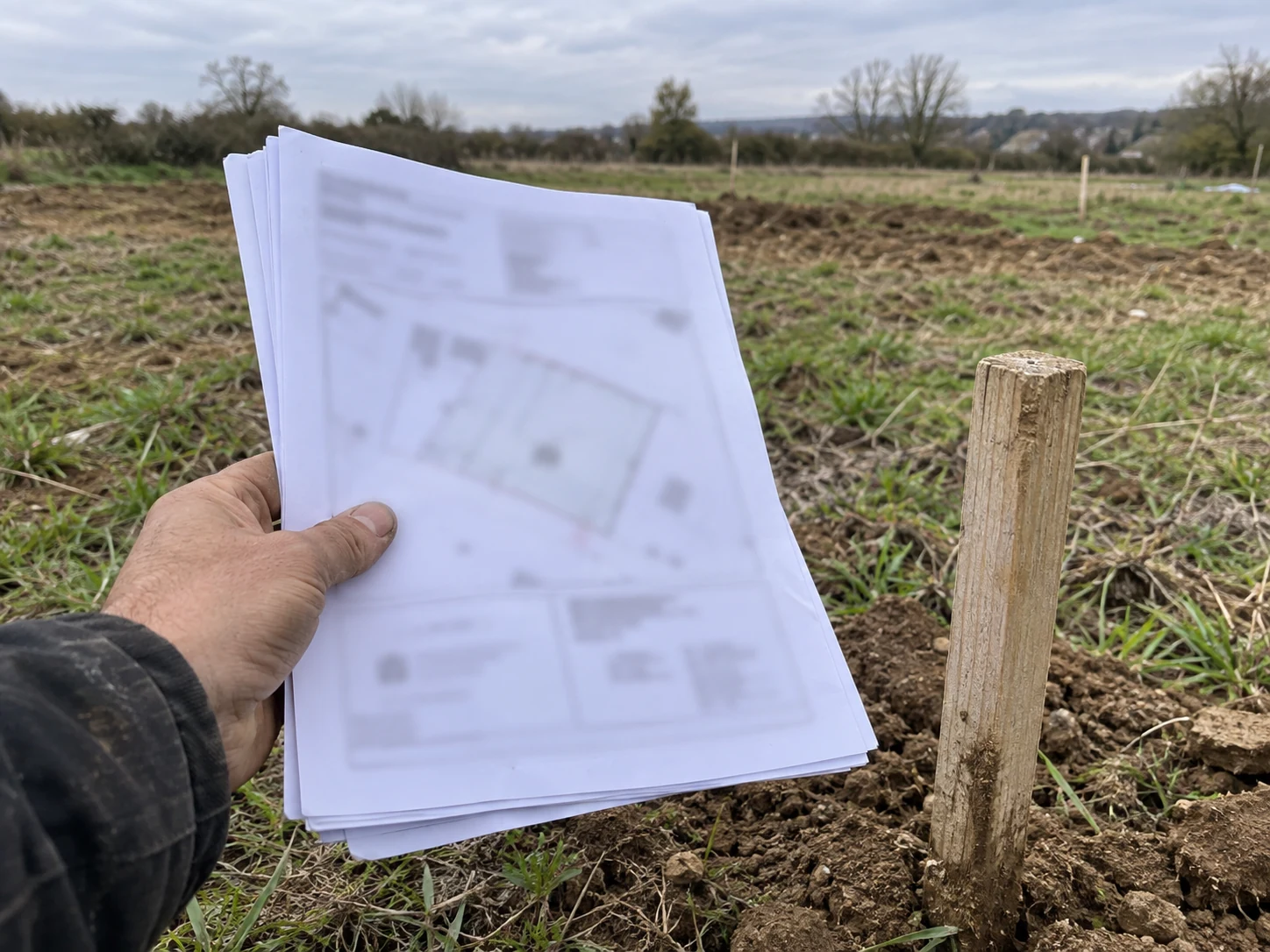 Main tenant des documents imprimés au-dessus d'un terrain à bâtir nu, près d'un piquet de bornage planté.