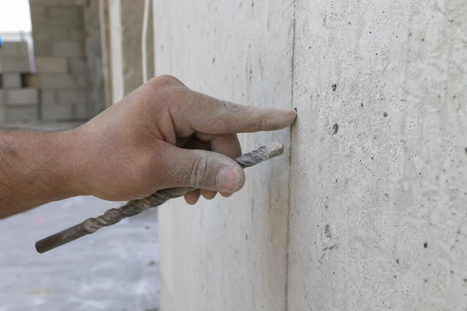Une main tenant une mèche approche la surface rugueuse d'un béton banché, gros plan sur le geste de diagnostic avant le perçage.