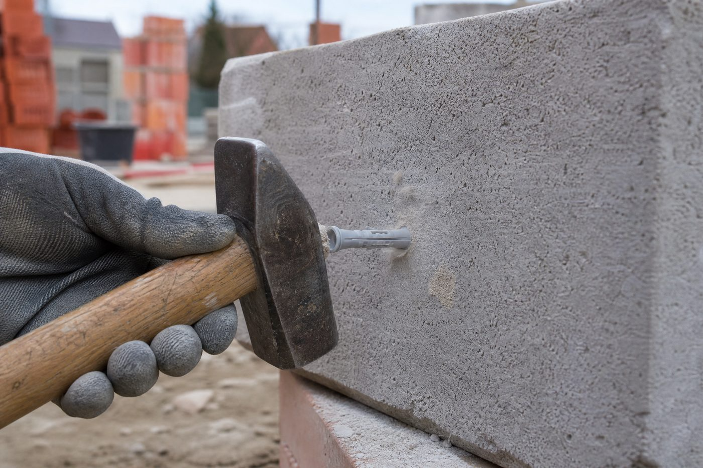 Main gantée enfonçant au marteau une cheville nylon grise dans un bloc de béton, en plein chantier.